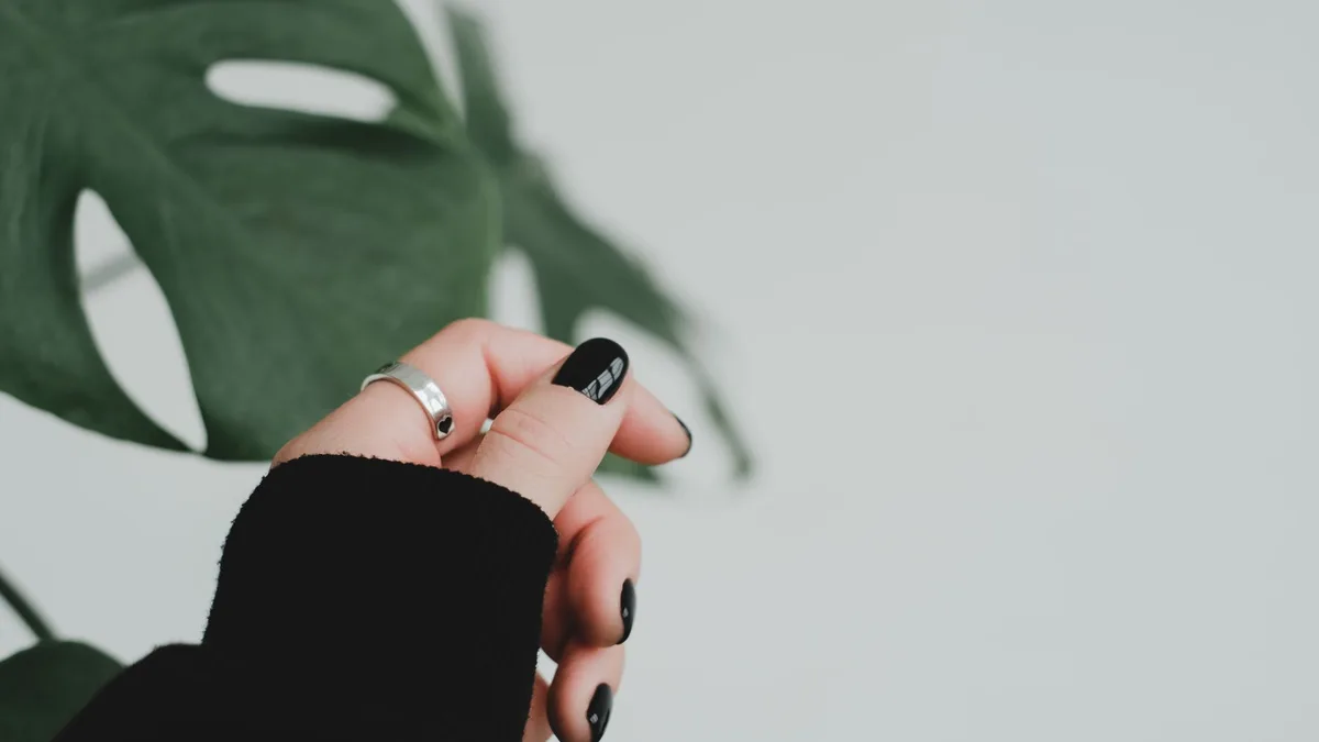 Close-up of a hand with black manicured nails and a silver ring against a green leaf background.