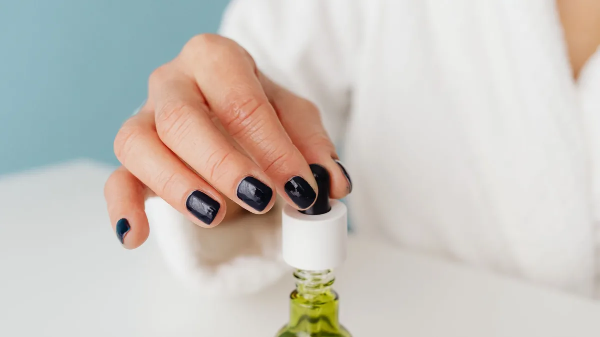 Hand holding a dropper over a glass bottle with green liquid, showcasing manicured nails.